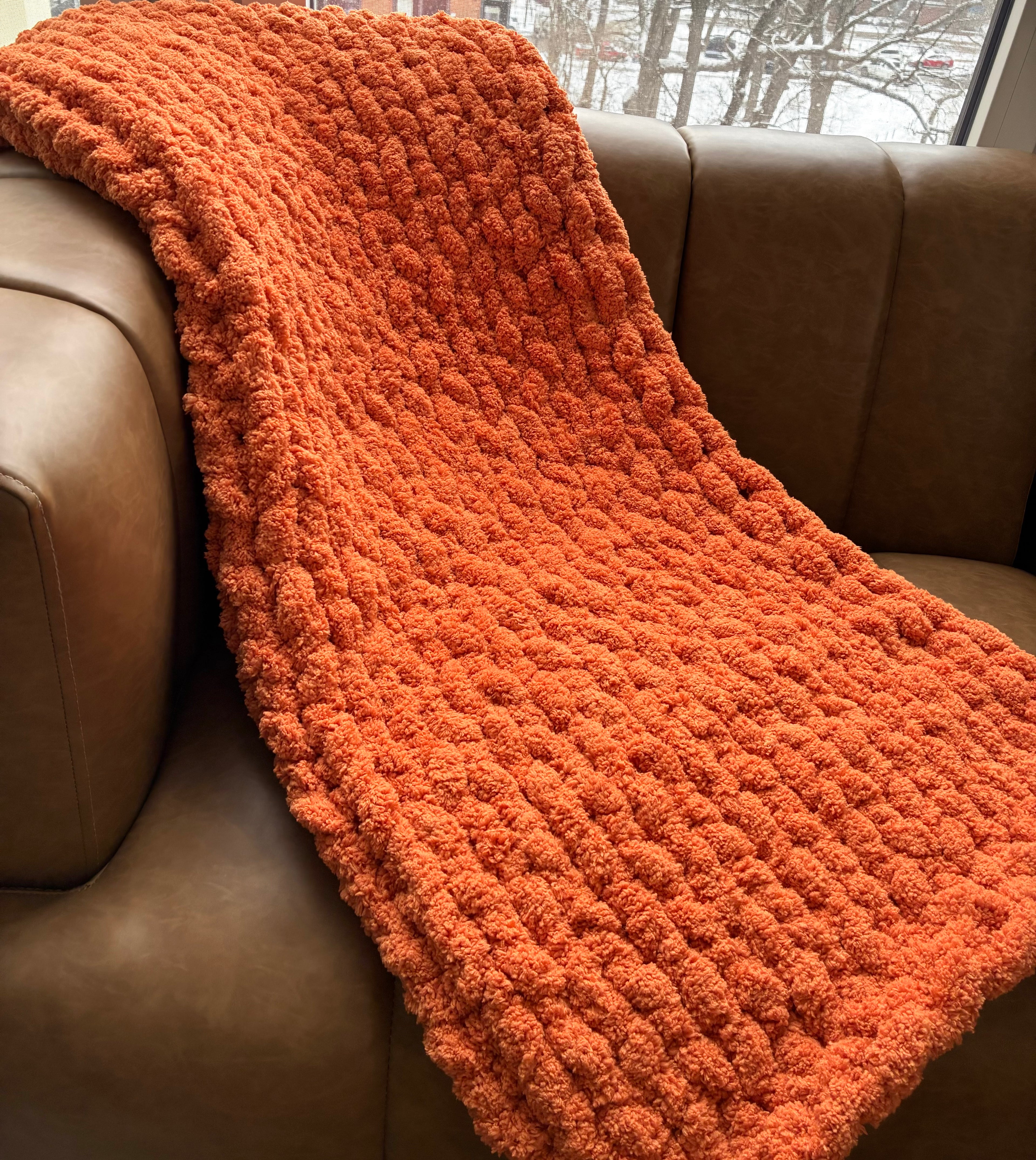 Orange textured blanket draped over a brown leather couch with a snowy outdoor scene in the background.